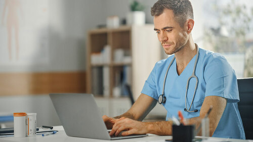 Experienced Male Nurse Wearing Blue Uniform Working on Laptop Co