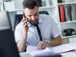 A man is sitting in the office, working with documents and talking on the phone.