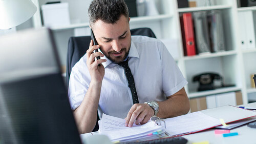 A man is sitting in the office, working with documents and talking on the phone.