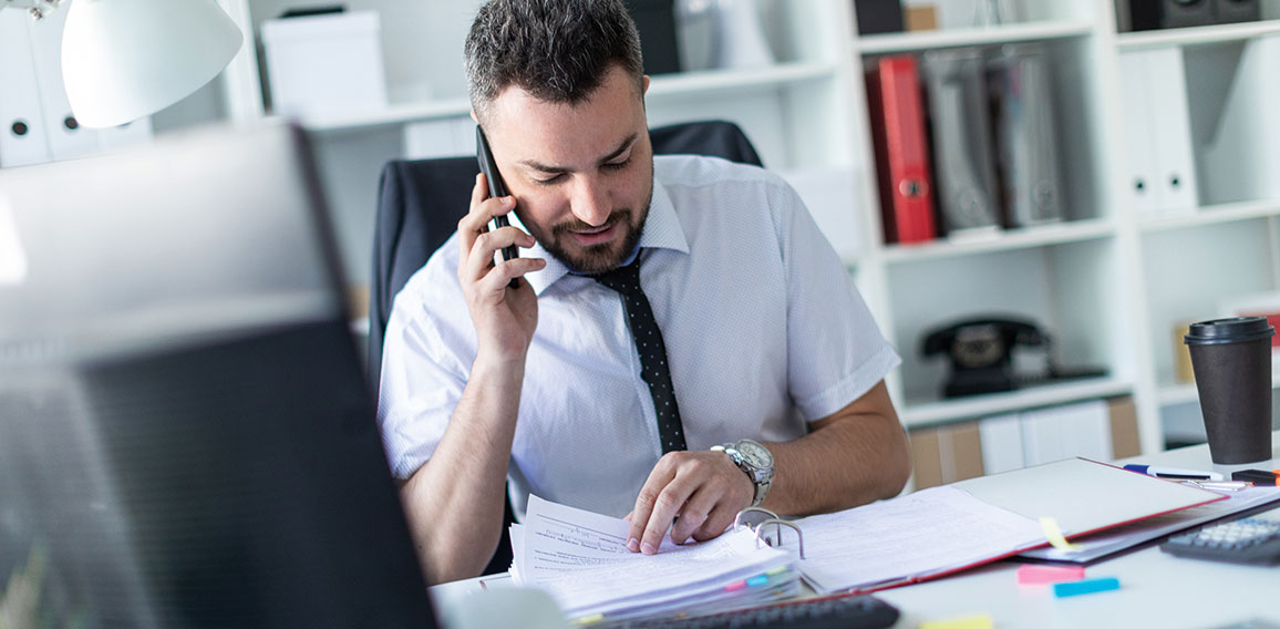 A man is sitting in the office, working with documents and talking on the phone.
