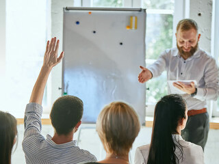 Male speaker giving talk at business meeting. Audience in conference hall. Rear view of businessman raising hand for speaker's question.