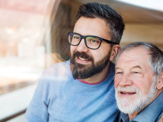 Adult son and senior father indoors at home, looking out. Shot through glass.