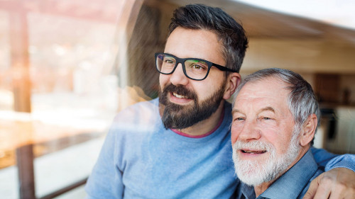Adult son and senior father indoors at home, looking out. Shot through glass.