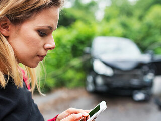 A young woman with smartphone by the damaged car after a car accident, text messaging.