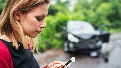 A young woman with smartphone by the damaged car after a car accident, text messaging.