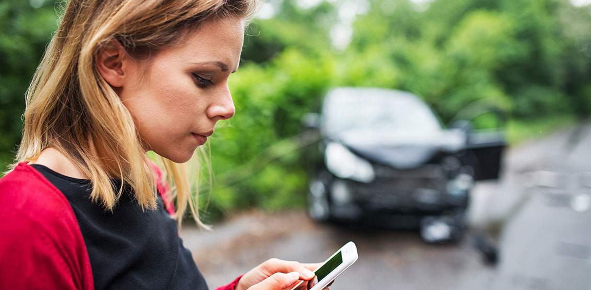 A young woman with smartphone by the damaged car after a car accident, text messaging.