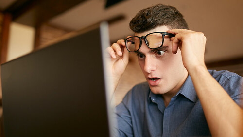 Shocked freelancer hipster man looks to laptop screen and can not believe unpleasant news. Pop-eyed frightened businessman trader raises one's glasses above his eyes. Trader monitoring stock exchange