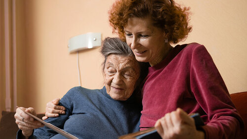 Senior woman and her adult daughter looking at photo album together on couch in living room, talking joyful discussing memories. Weekend with parents, family day, thanksgiving, mom's holiday