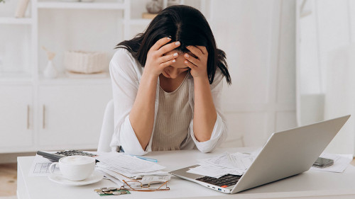Desperate young woman holds head in hands, feeling overwhelmed by financial paperwork at white office desk, struggling with bills, taxes, and credit management, bankruptcy concept, mental health