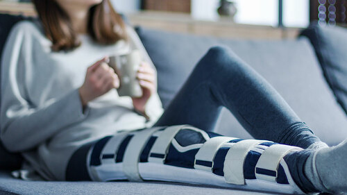 Smiling brunette girl with broken leg is sitting on couch sofa,